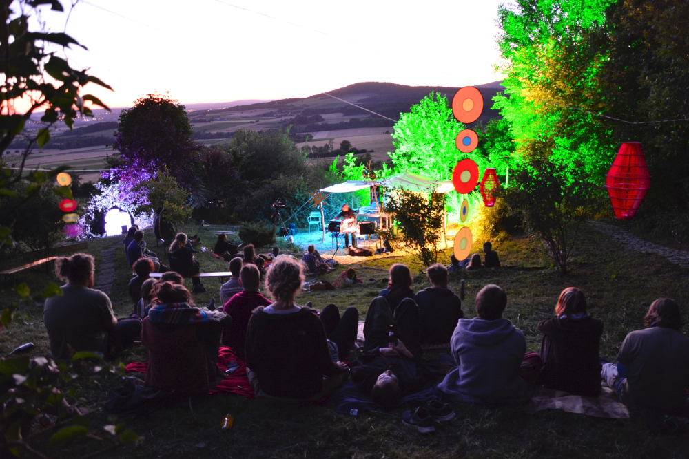 Publikum lauscht bei Abenddämmerung auf der Wiese vor der Bühne